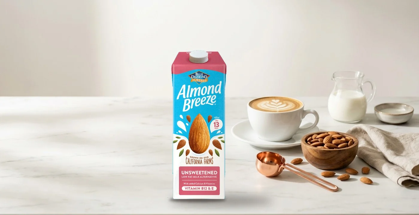 Almond Breeze Unsweetened carton on a white marble counter beside a flat white, a bowl of almonds and a copper scoop