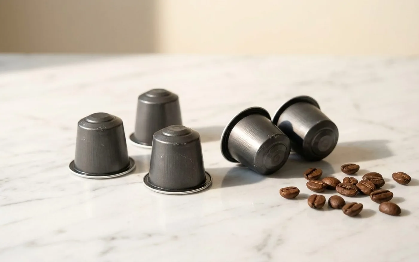 Five aluminium coffee capsules arranged on a white marble surface with scattered coffee beans
