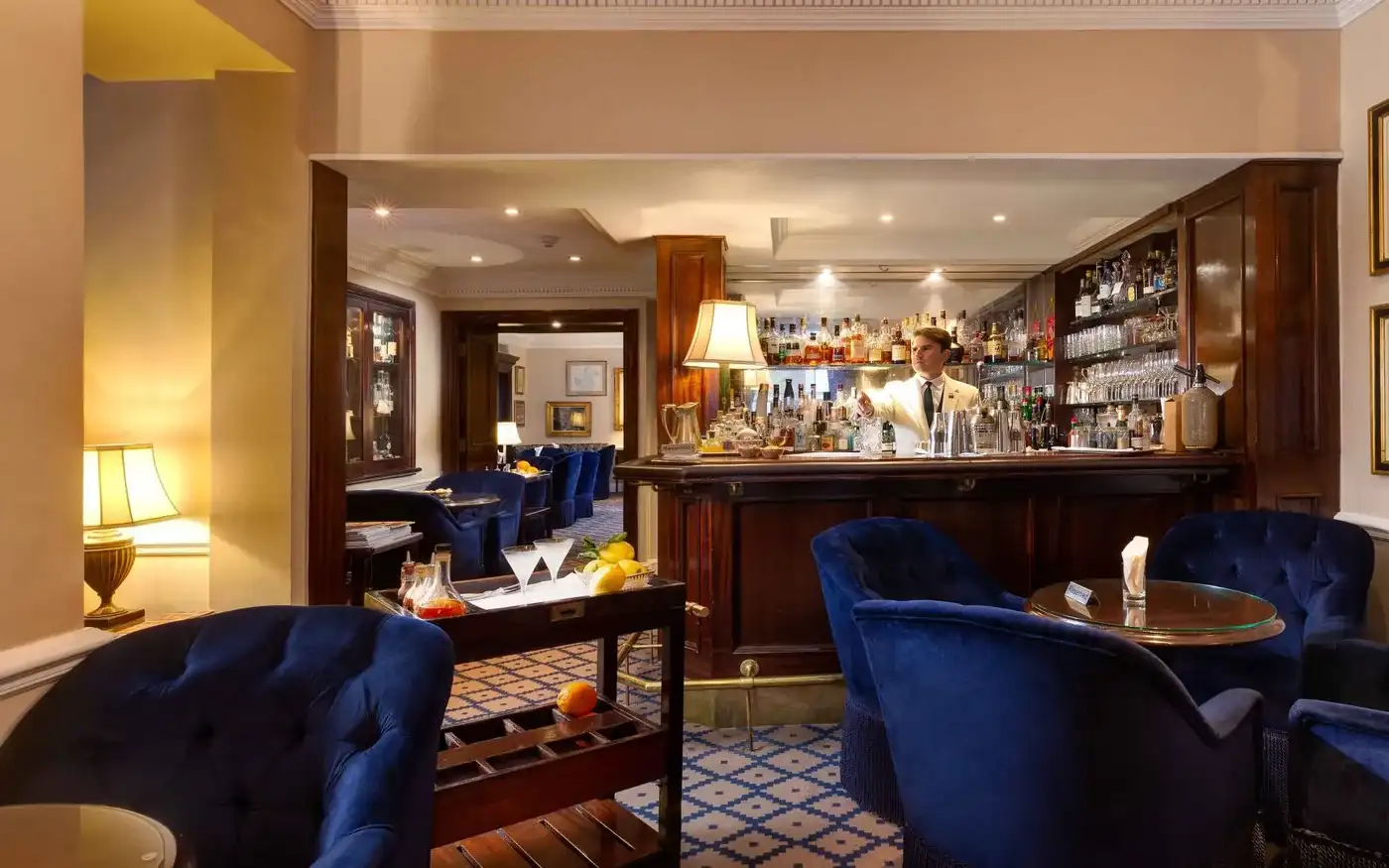 Bartender behind the bar with blue velvet armchairs at Dukes Bar in St James's, London