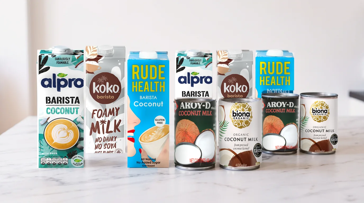 Five coconut milk products for coffee lined up on a kitchen counter, including barista cartons and tinned options