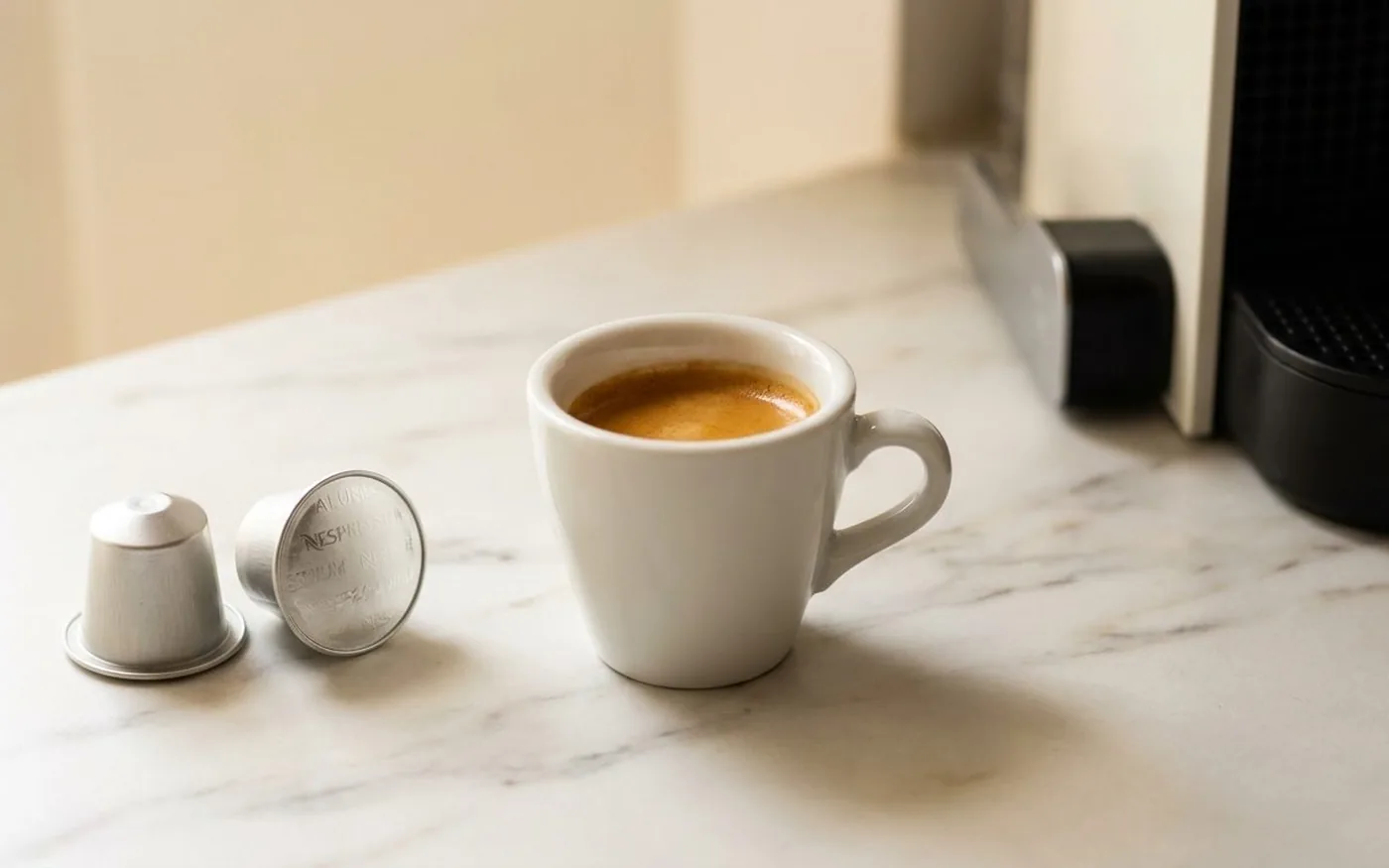 Freshly pulled espresso shot in a small white ceramic cup on a white marble countertop, with two aluminium Nespresso-compatible coffee capsules resting nearby and a Nespresso machine partially visible in the background