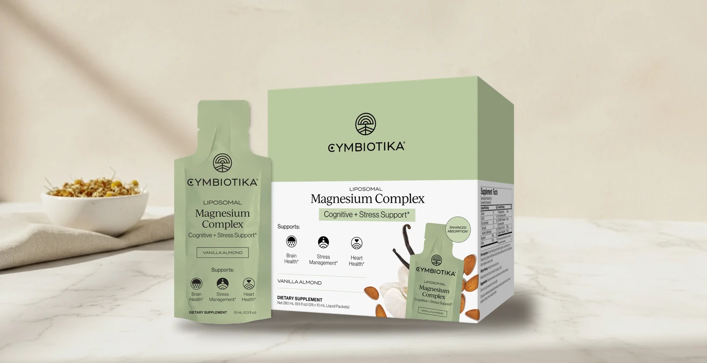 Cymbiotika Liposomal Magnesium Complex vanilla almond pouch and retail box on a white marble counter beside a bowl of dried chamomile and a linen cloth, against a warm cream wall
