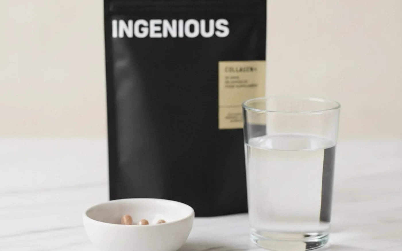 Three Ingenious Beauty collagen capsules in a ceramic bowl beside a glass of water and the COLLAGEN+ pouch