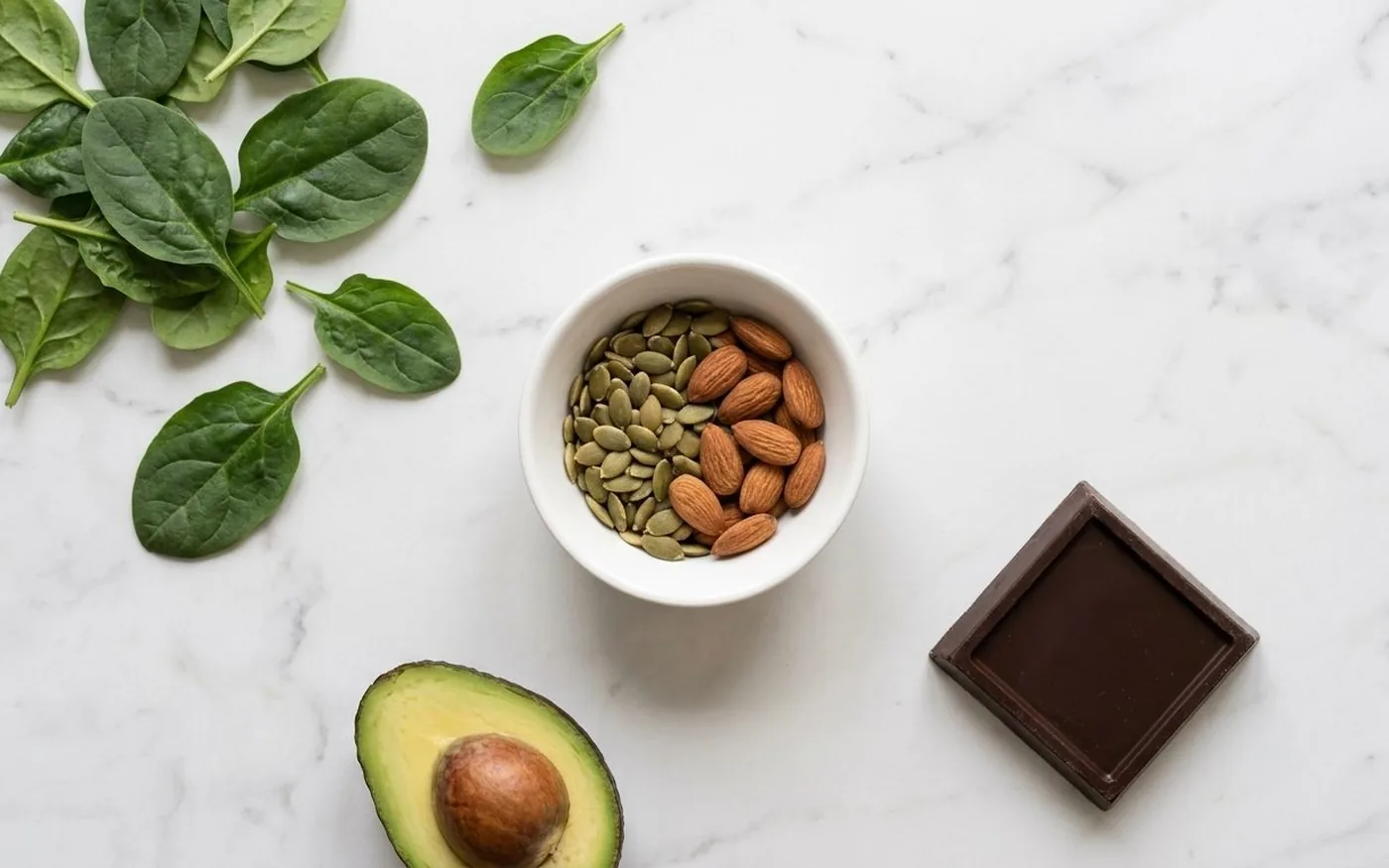 Overhead flat lay of magnesium-rich foods including fresh spinach, pumpkin seeds, almonds, dark chocolate, and avocado on white marble