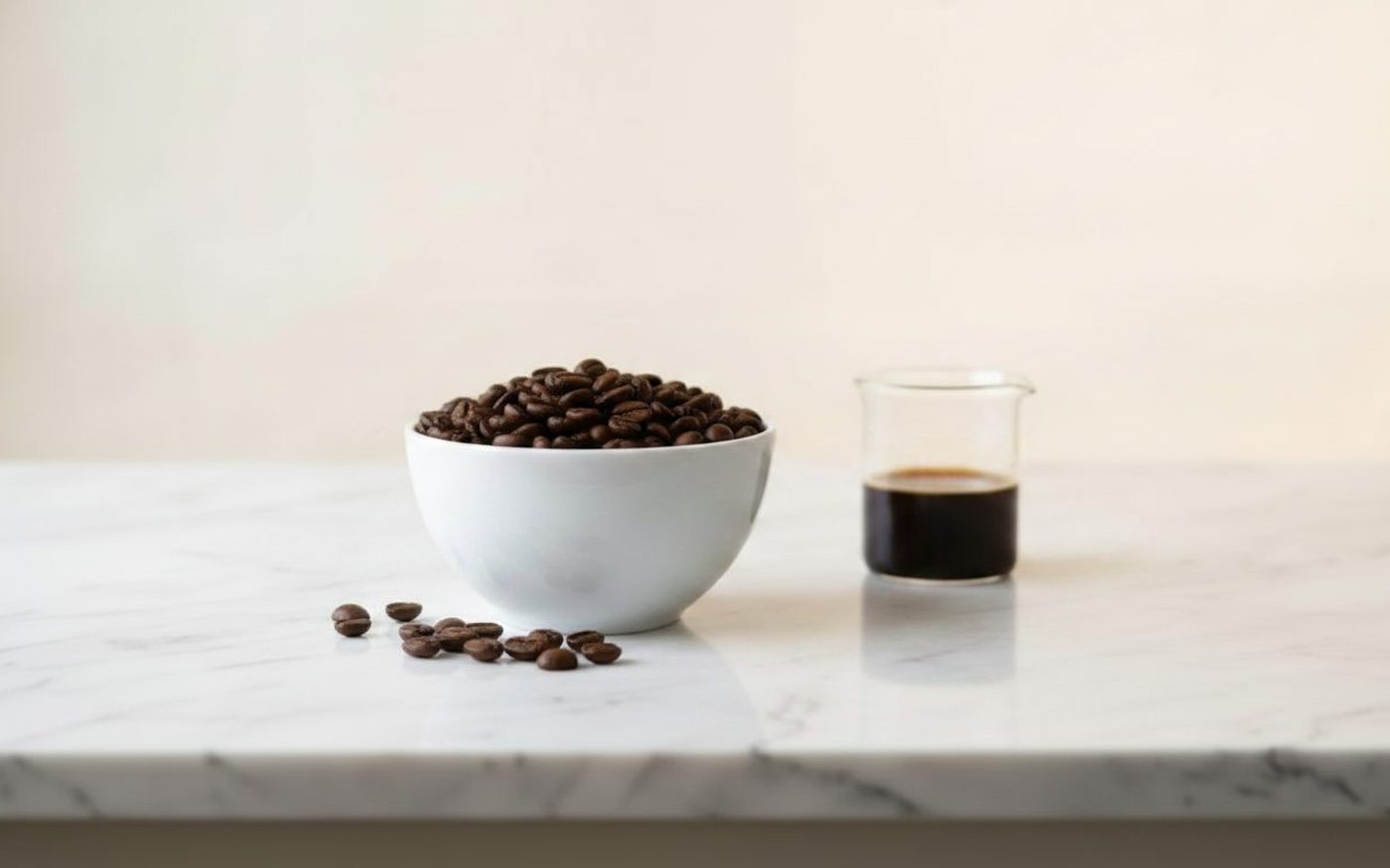 Roasted coffee beans in white bowl beside glass beaker on white marble