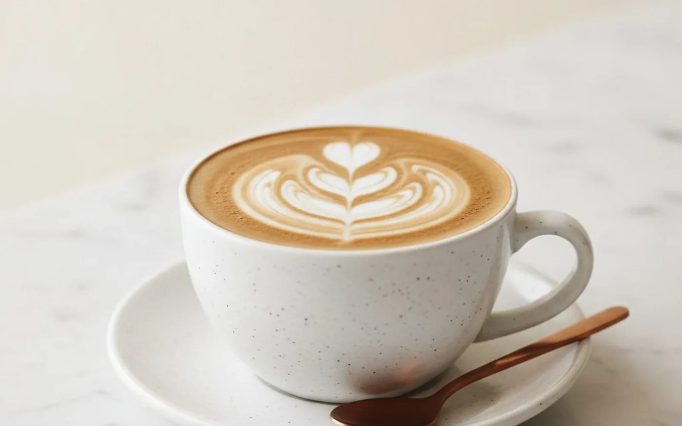 Flat white with heart-shaped latte art in a speckled cup, copper spoon on the saucer