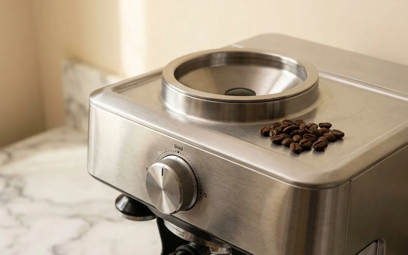 Close-up of the Sage Barista Express integrated conical burr grinder module on top of the machine body, with coffee beans beside the hopper on a marble countertop