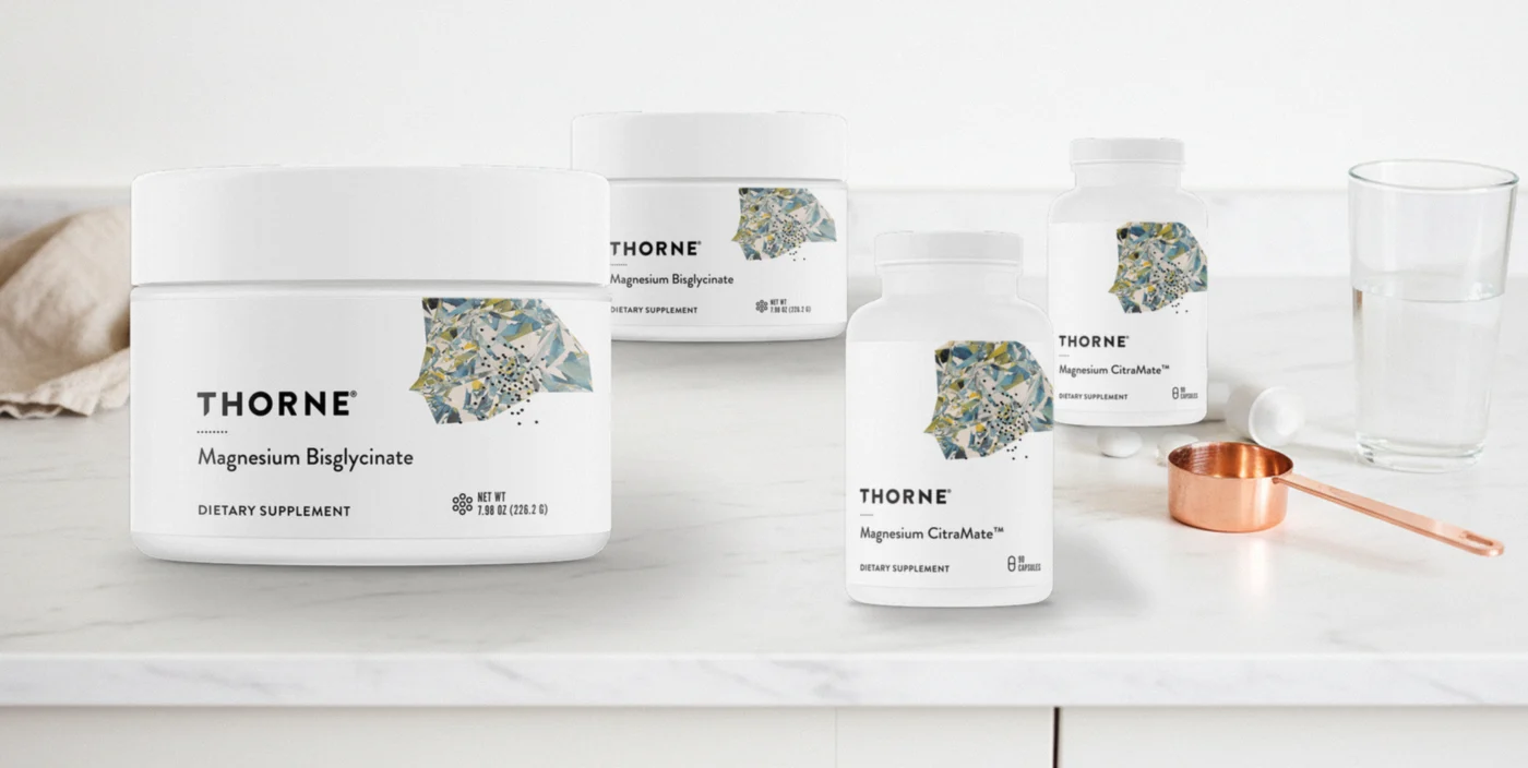 Thorne Magnesium Bisglycinate powder tubs and Magnesium CitraMate capsule bottles with legible labels on white marble beside a linen cloth, copper scoop and glass of water